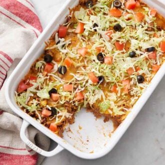 An overhead view of a baking dish of taco casserole with a slice taken out. Lettuce, tomatoes, and black olives on top.