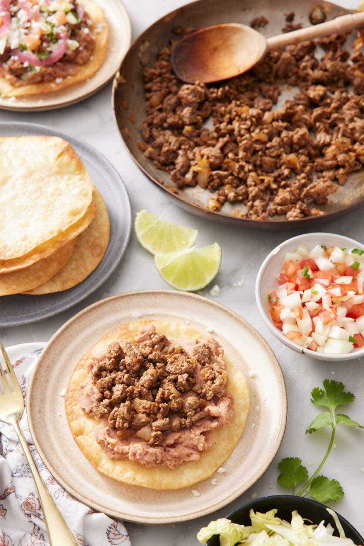 A plate with a tostada with no toppings. A plate of fried tortillas, bowl of pico de gallo, lime wedges, cooked beef, and another tostada in the background.