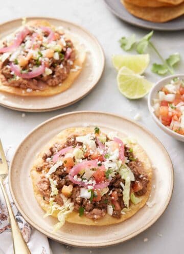 A plate with a tostada with a second plate in the background along with cut limes, cilantro, and a bowl of pico de gallo.