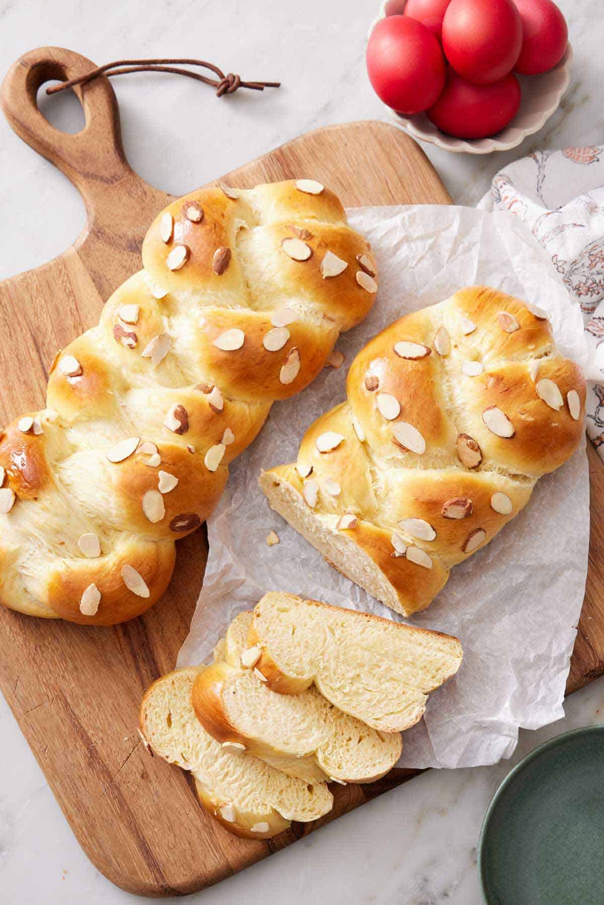 Overhead view of a serving board with parchment paper and two loaves of tsoureki, one with three slices cut.