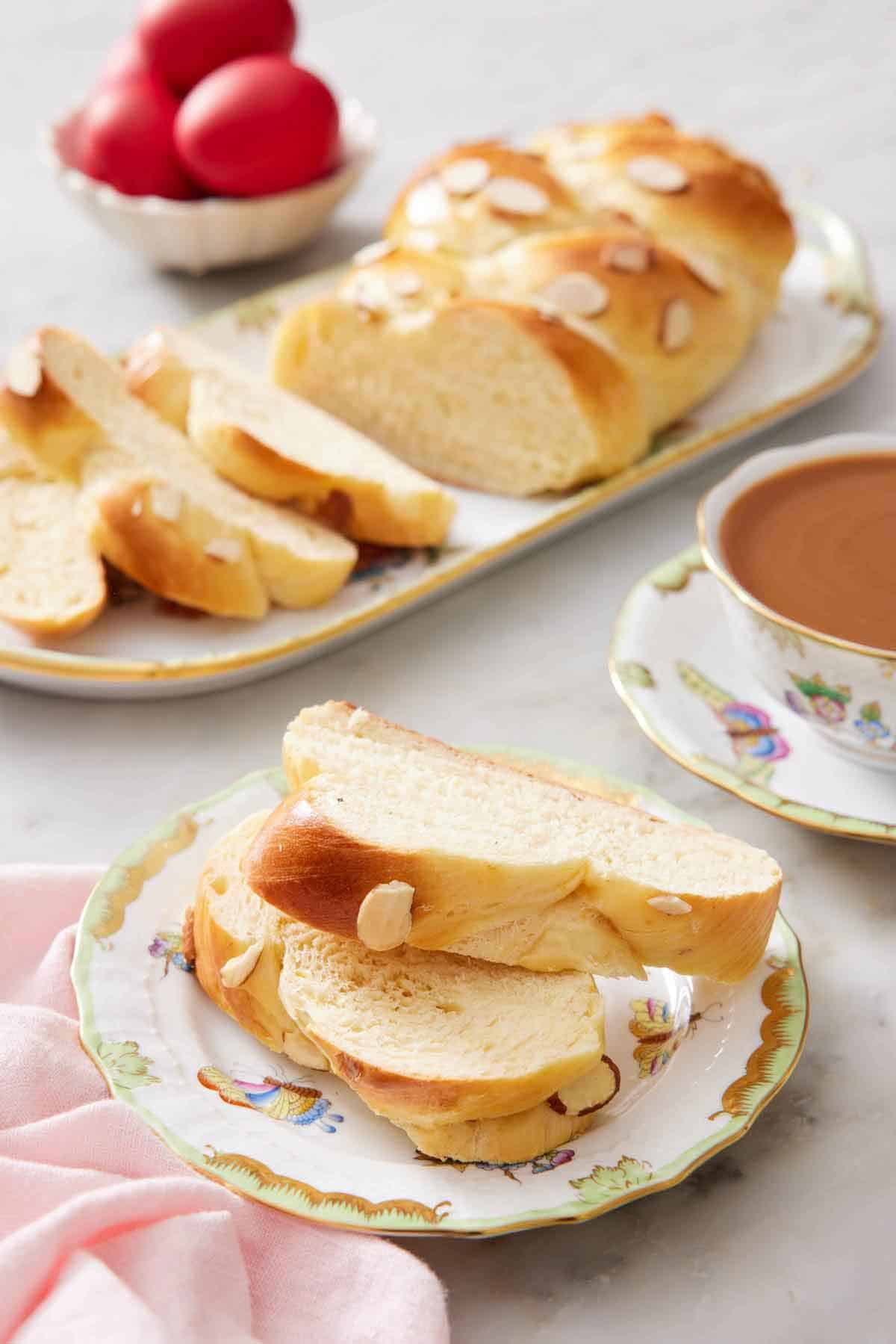 A plate with two slices of tsoureki with a coffee in the back along with a platter with the rest of the bread.