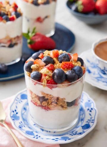 A glass of yogurt parfait on a plate with a platter in the background with two more parfaits.