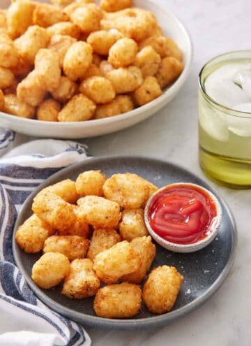 A plate of air fryer tater tots with a bowl of ketchup. A drink in the background along with a large bowl of more tater tots.