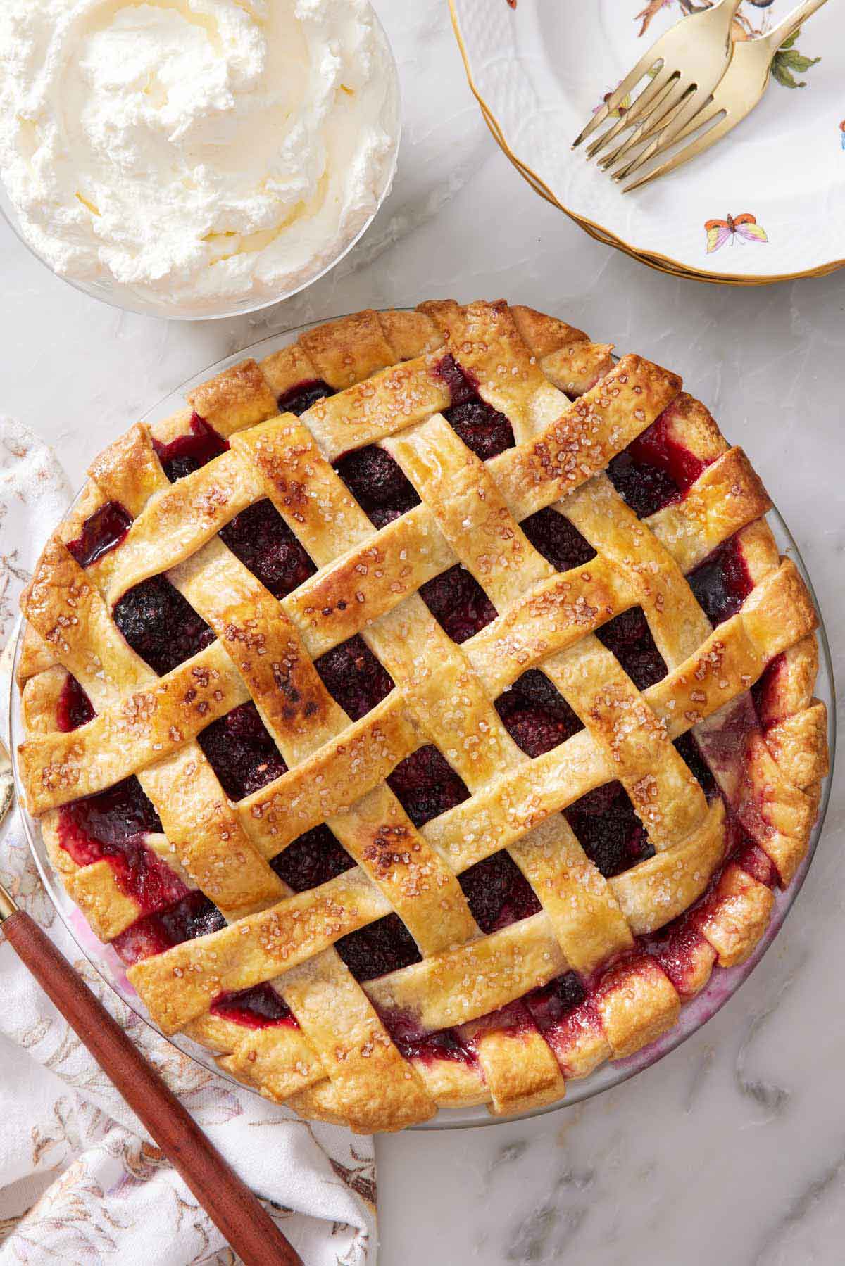 Overhead view of a blackberry pie. Whipped cream on the side along with some forks on a plate.