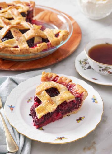 A plate with a slice of blackberry pie with the rest of the pie in the background along with a mug of tea.