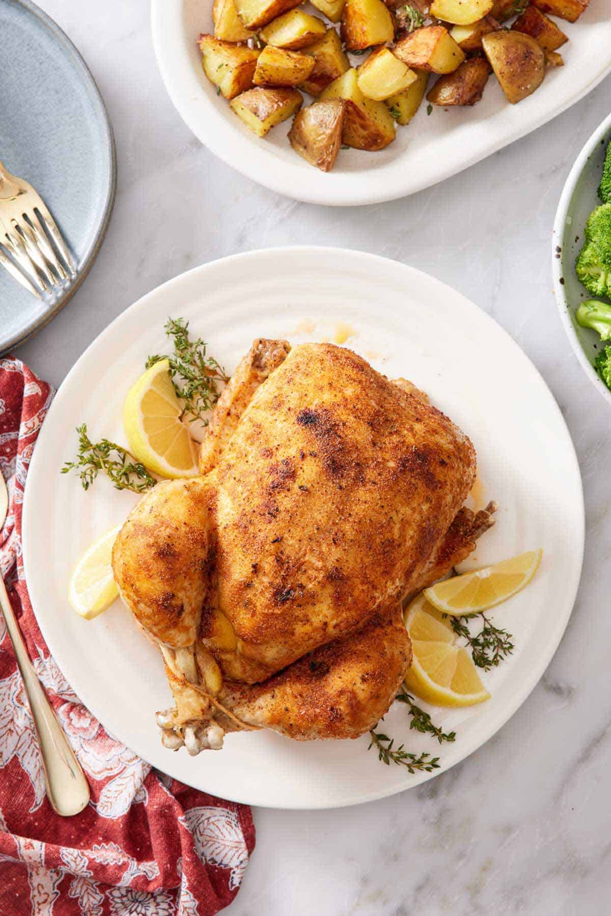 Overhead view of an instant pot whole chicken over fresh thyme and lemon wedges on a white plate. Roast potatoes on the side.