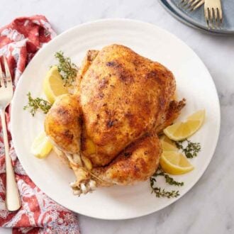 Overhead view of a white plate with an instant pot whole chicken over fresh thyme and lemon wedges. A fork on the side with a napkin.