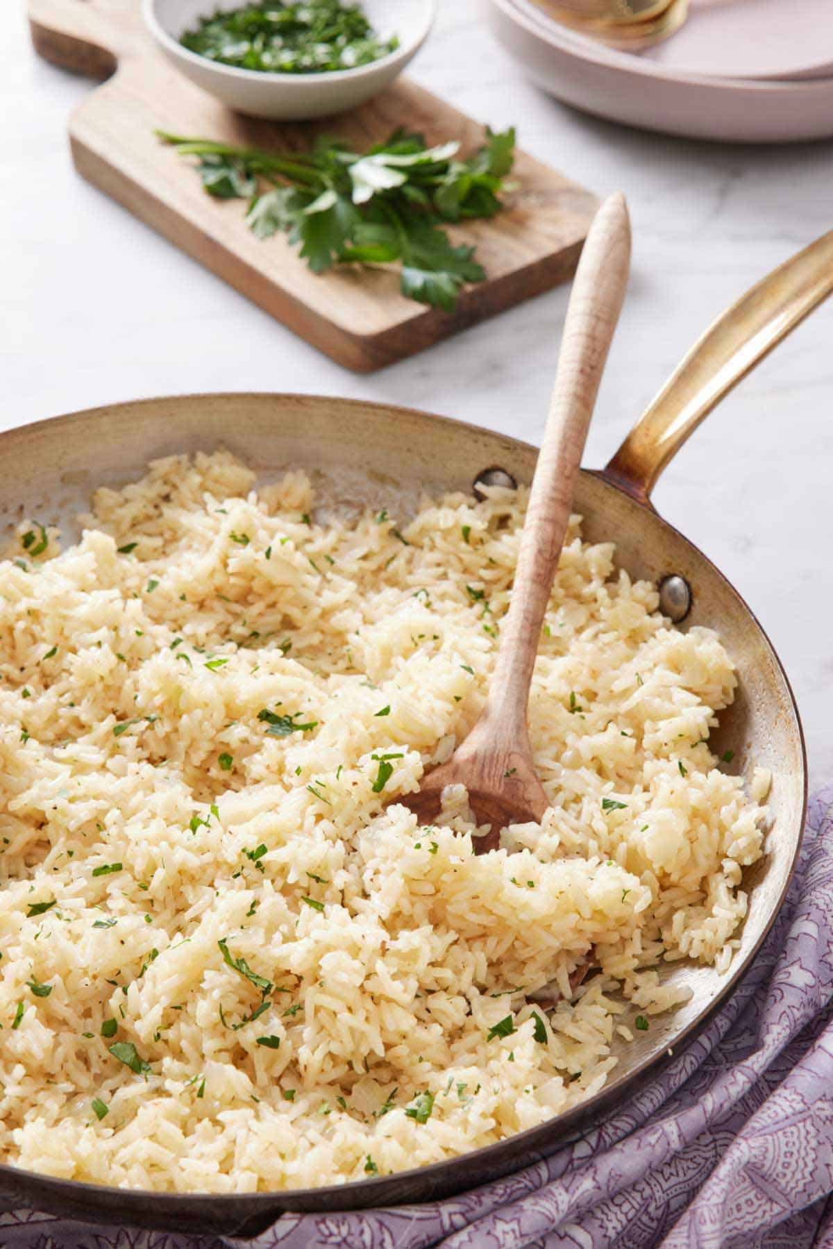 A skillet of rice pilaf with a wooden spoon tucked in. Parsley in the background on a cutting board.