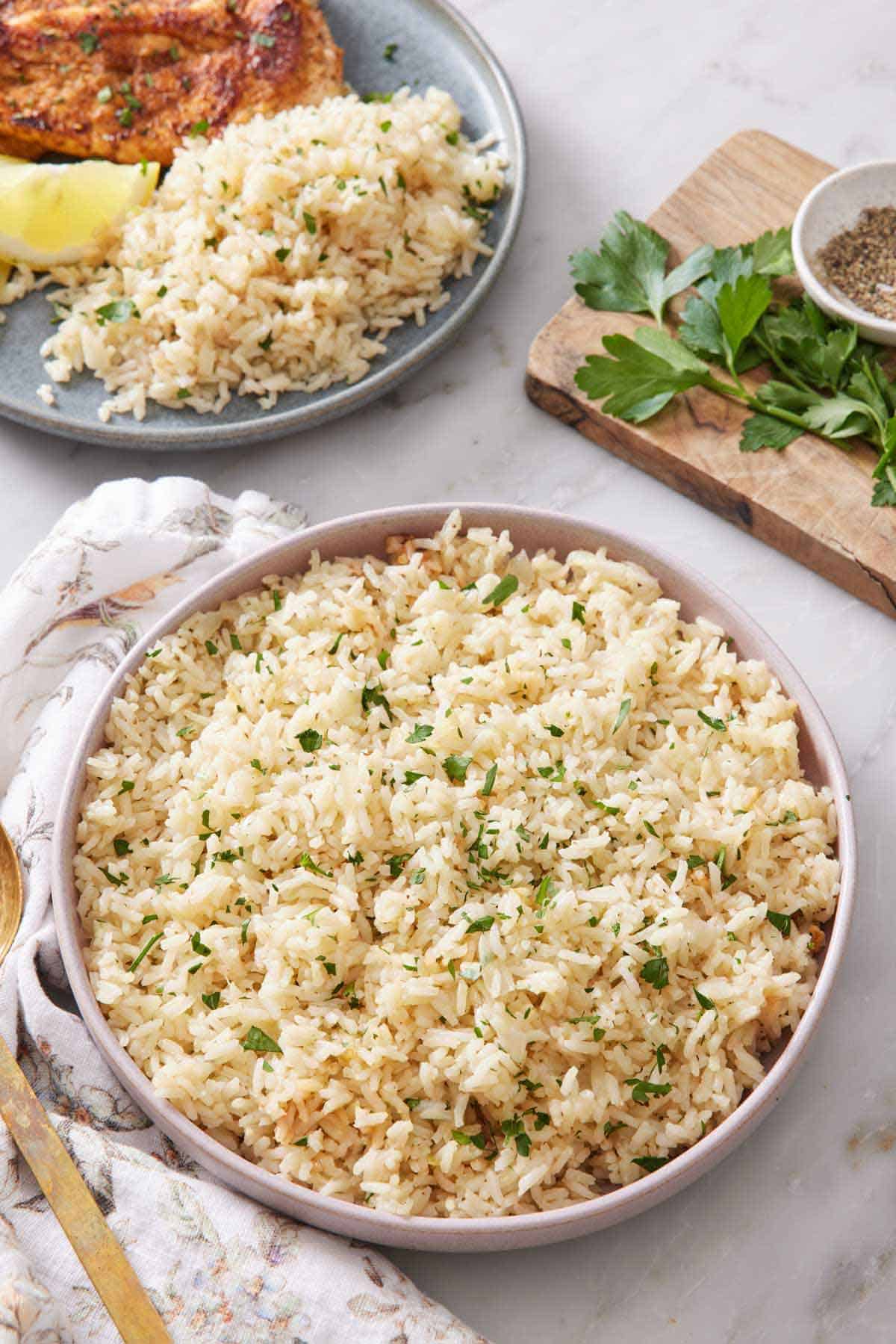 A large plate of rice pilaf with chopped parsley garnish. Another plate in the background with rice pilaf along with a lemon wedge and meat.