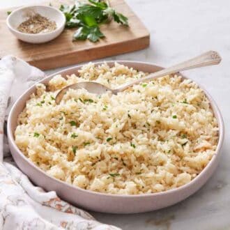 A plate of rice pilaf with a spoon tucked in. A wooden board with parsley and bowl of pepper in the back.