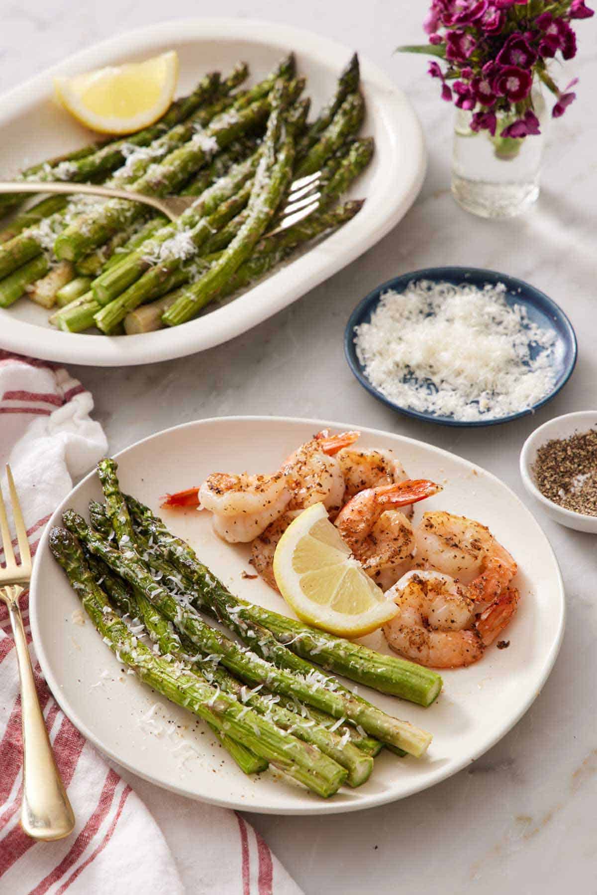 A plate of shrimp with air fryer asparagus and a lemon wedge. A platter of more air fryer asparagus in the back along with a bowl of parmesan.