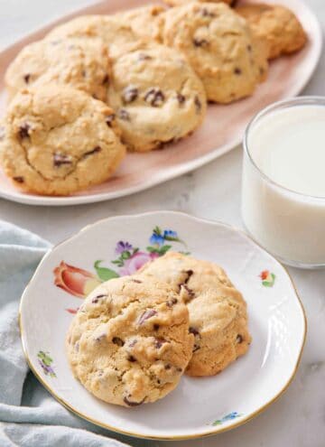 A plate with two air fryer cookies with a glass of milk and a platter of more cookies in the back.