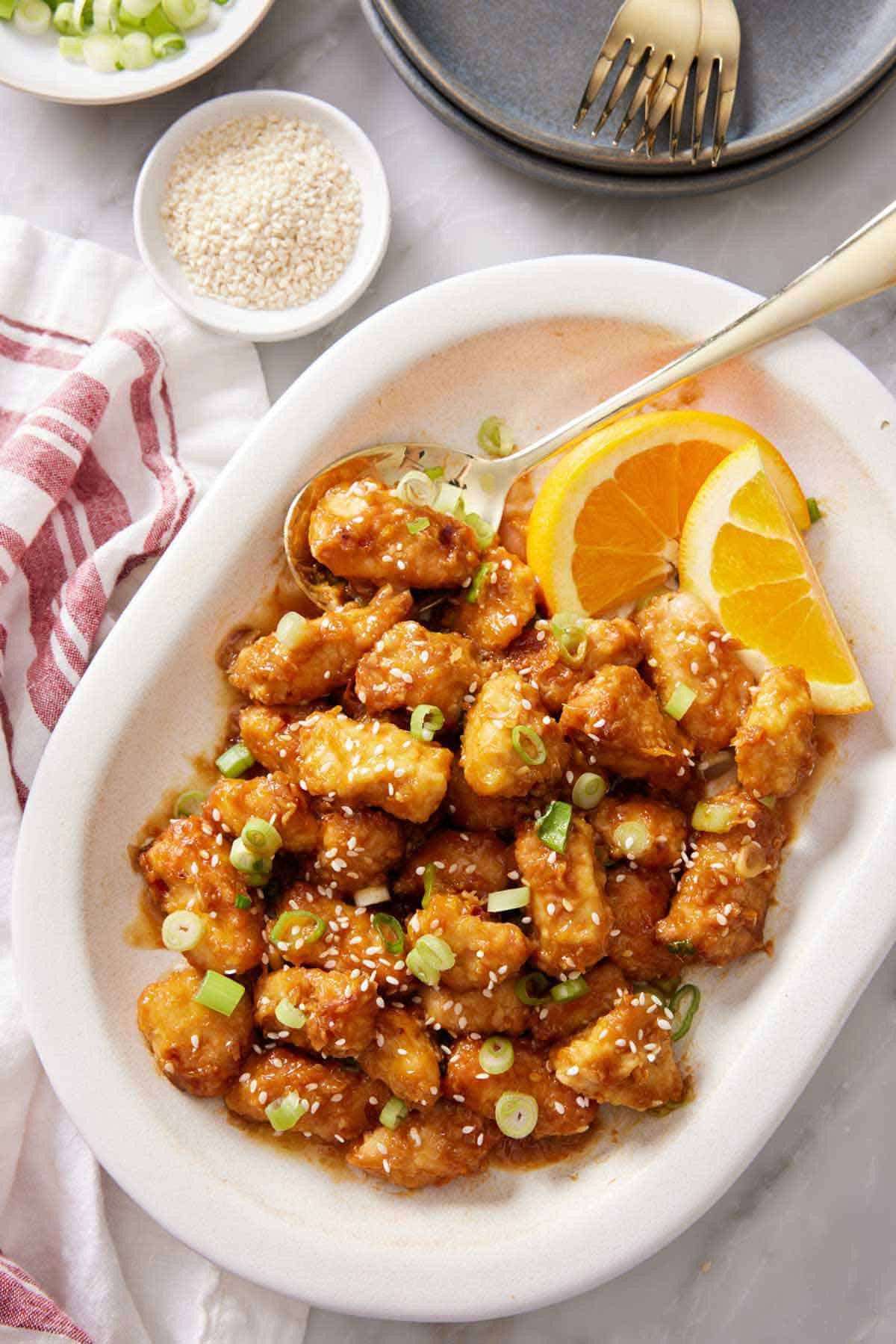 Overhead view of a platter of air fryer orange chicken topped with green onions and sesame seeds with some orange wedges.