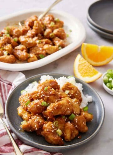 A plate with air fryer orange chicken over rice. A platter in the background with more chicken. Orange wedges on the side.