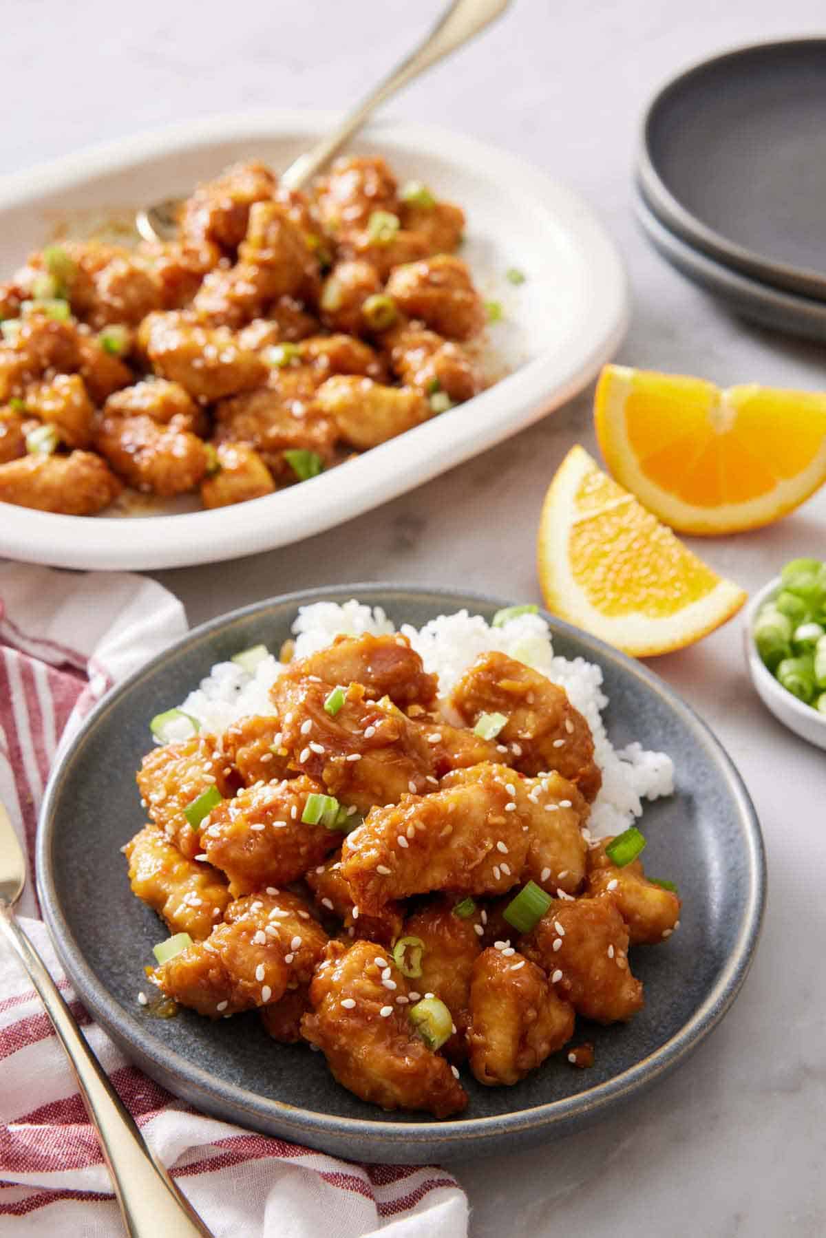 A plate with air fryer orange chicken over rice. A platter in the background with more chicken. Orange wedges on the side.