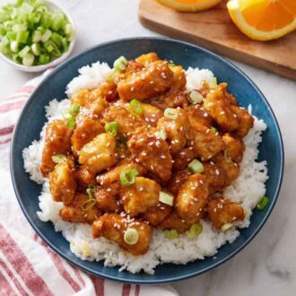 An overhead view of a plate of rice with air fryer orange chicken on top. A bowl of green onions and some cut orange wedges in the back.
