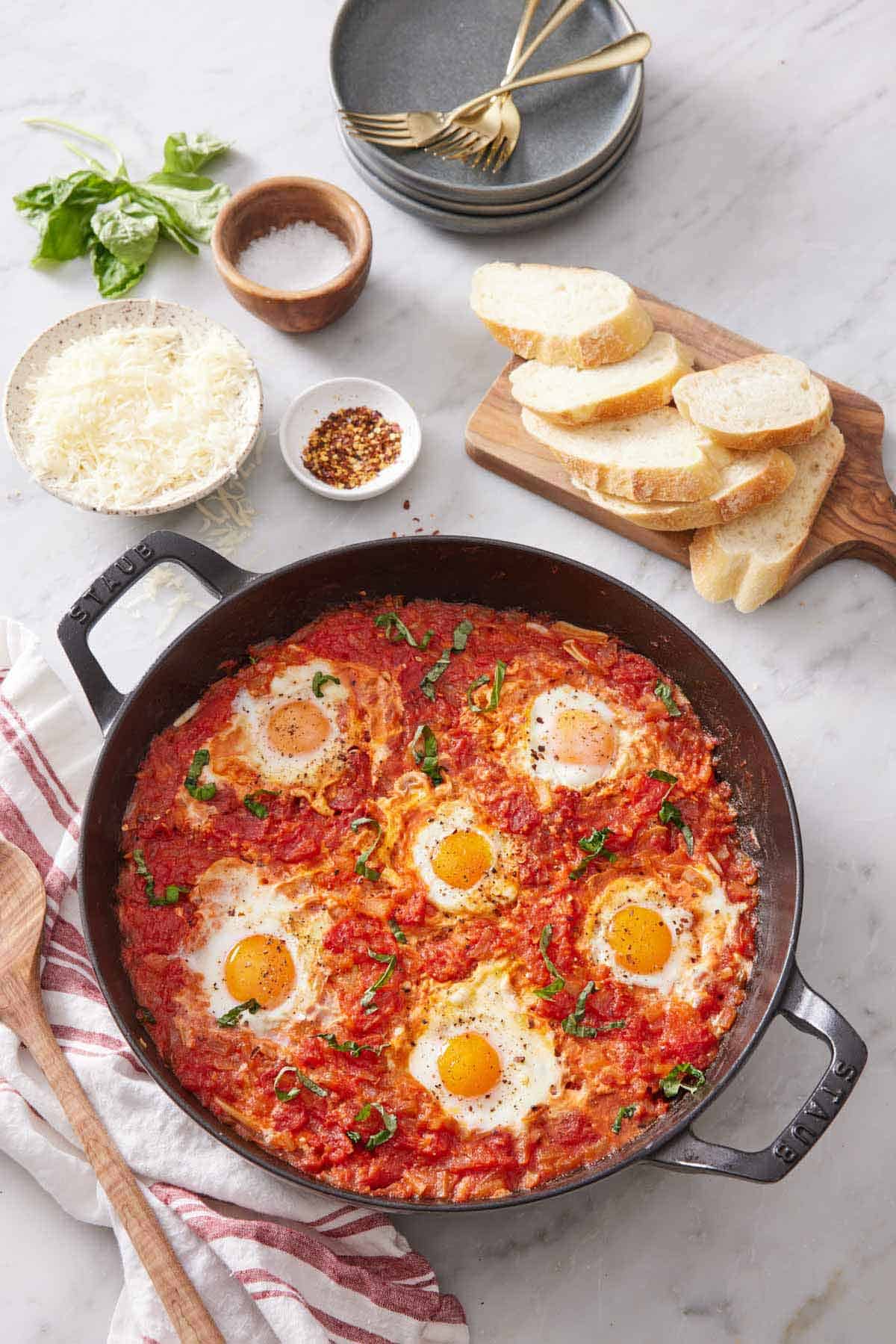 Overhead view of a skillet of eggs in purgatory. A board with sliced bread, bowl of shredded cheese, basil, salt, and red pepper flakes.