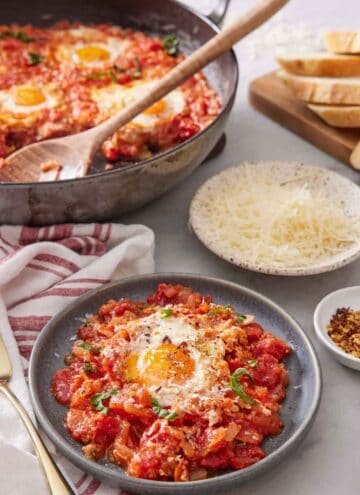 A plate of eggs in purgatory with a skillet in the background with the rest of the eggs in purgatory along with a plate of shredded cheese.