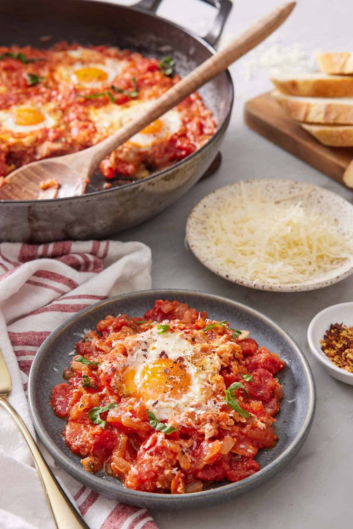 A plate of eggs in purgatory with a skillet in the background with the rest of the eggs in purgatory along with a plate of shredded cheese.