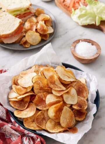 A plate lined with parchment paper topped with homemade potato chips. A plate with a cut sandwich with chips in the back along with a bowl of salt and sandwich ingredients.