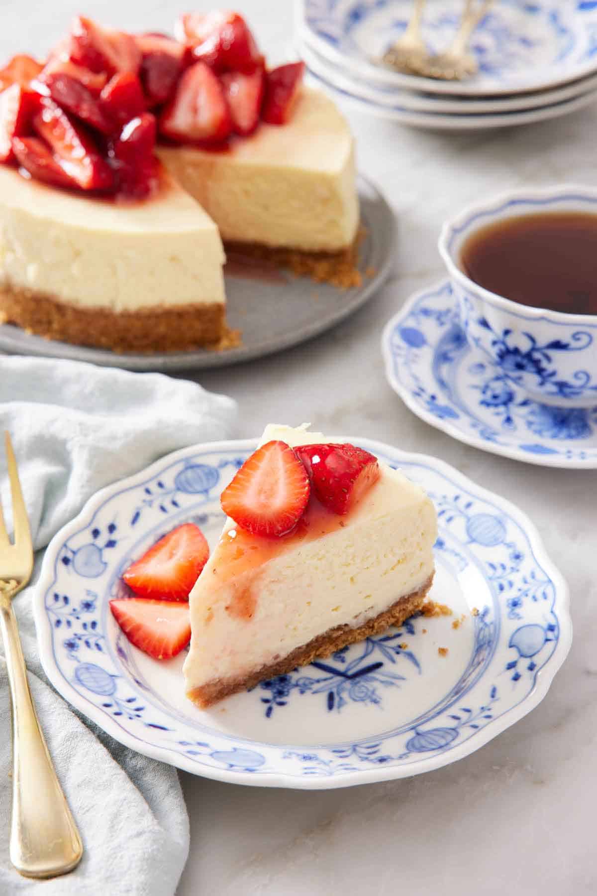 A plate with a slice of Instant Pot cheesecake topped with chopped strawberries. A mug of tea and the rest of the cheesecake in the background.