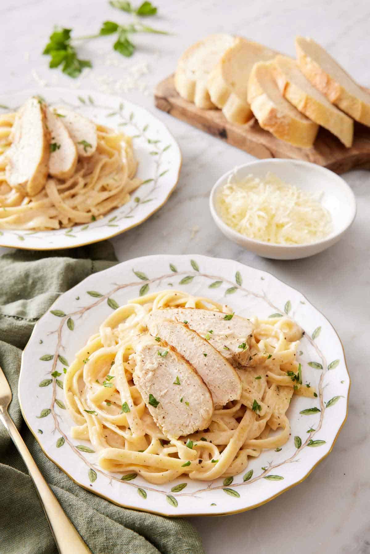 A plate with a serving of Instant Pot chicken alfredo with a second plate in the back along with a bowl of cheese and sliced bread.