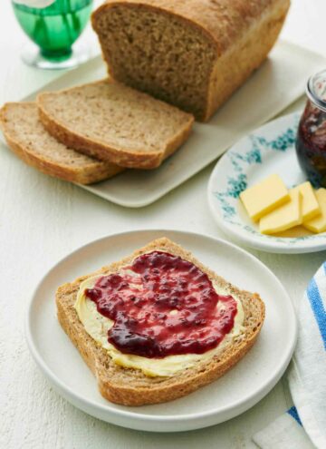A plate with a slice of rye bread with peanut butter and jam spread on top. Rest of the loaf in the background with two more slices cut.