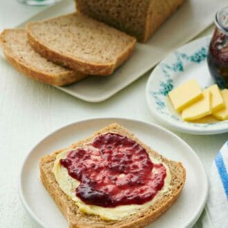 Pinterest graphic of a plate with a slice of rye bread with peanut butter and jam spread on top. Rest of the loaf in the background with two more slices cut.