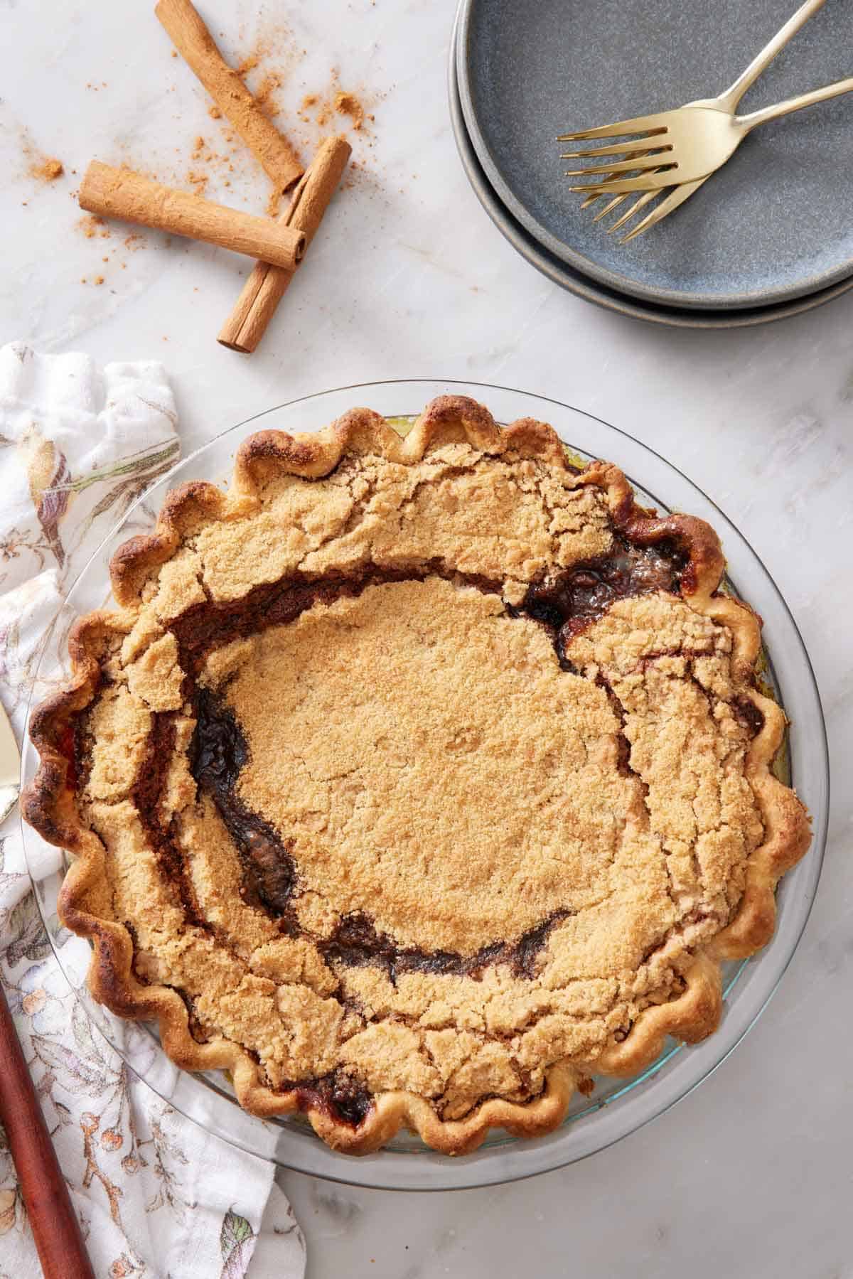 Overhead view of a shoofly pie in a glass pie dish. Plates, forks, and some cinnamon sticks on the side.