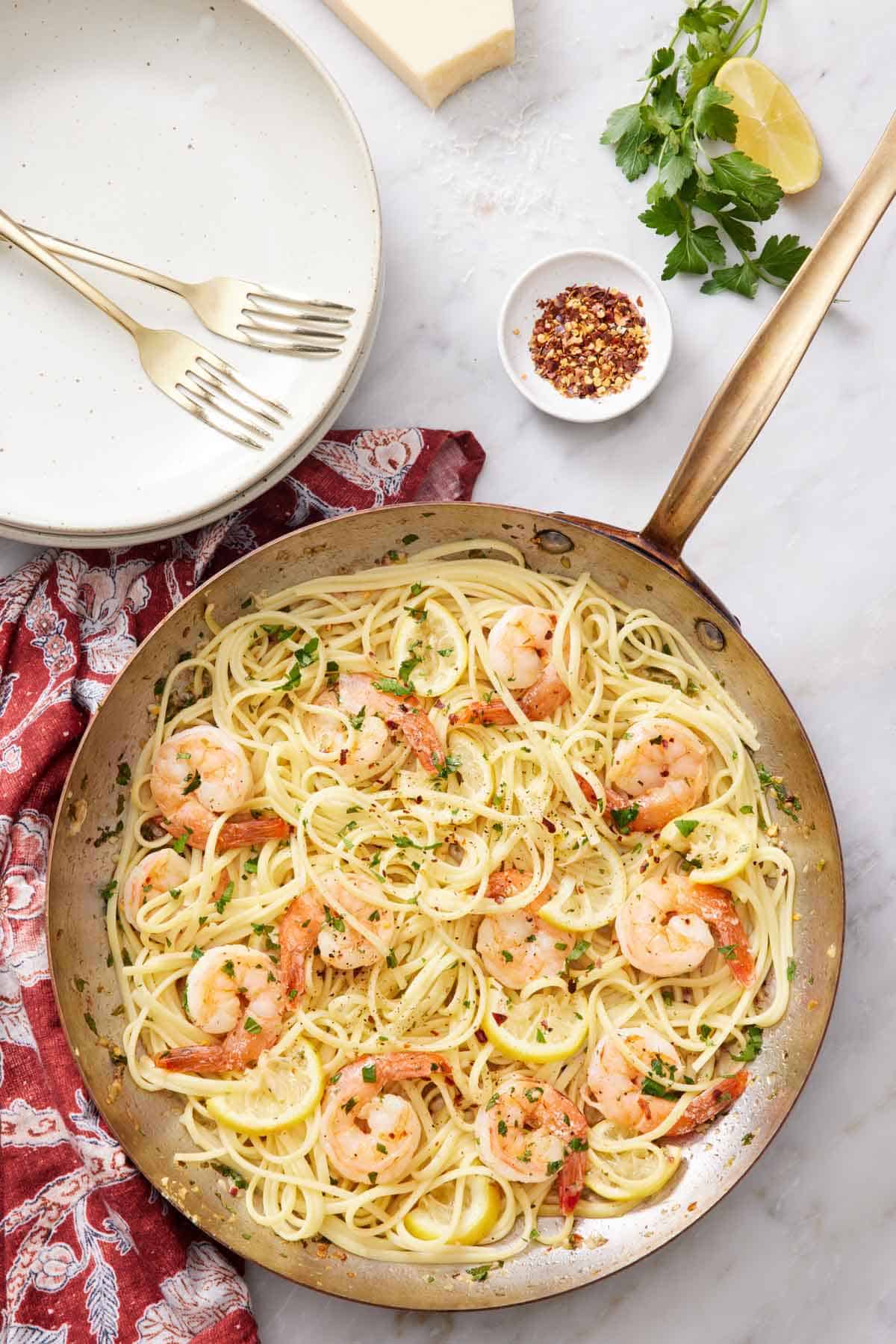 Overhead view of a skillet of shrimp linguine. Plates, forks, red pepper flakes, lemon wedge, and parsley off to the side.