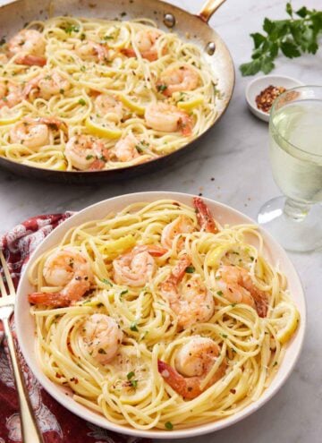 A plate with shrimp linguine with a glass of wine and skillet with more shrimp linguine in the back.