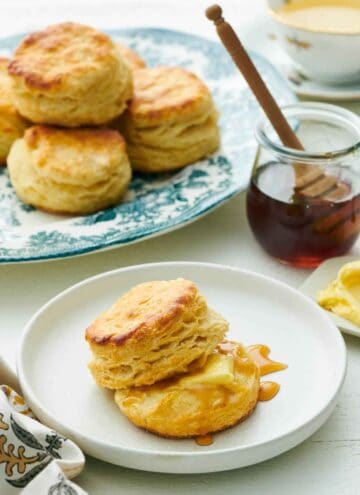 A plate with buttermilk biscuits with some honey and butter. A jar of honey and platter of biscuits in the background.