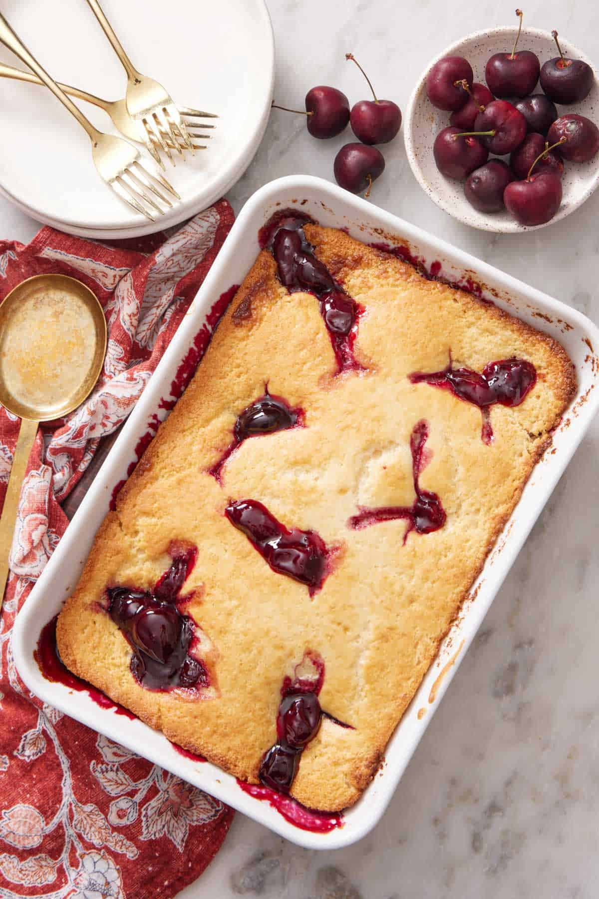 Overhead view of a baking dish of cherry cobbler with a bowl of cherries, stack of plates, and three forks on the side.