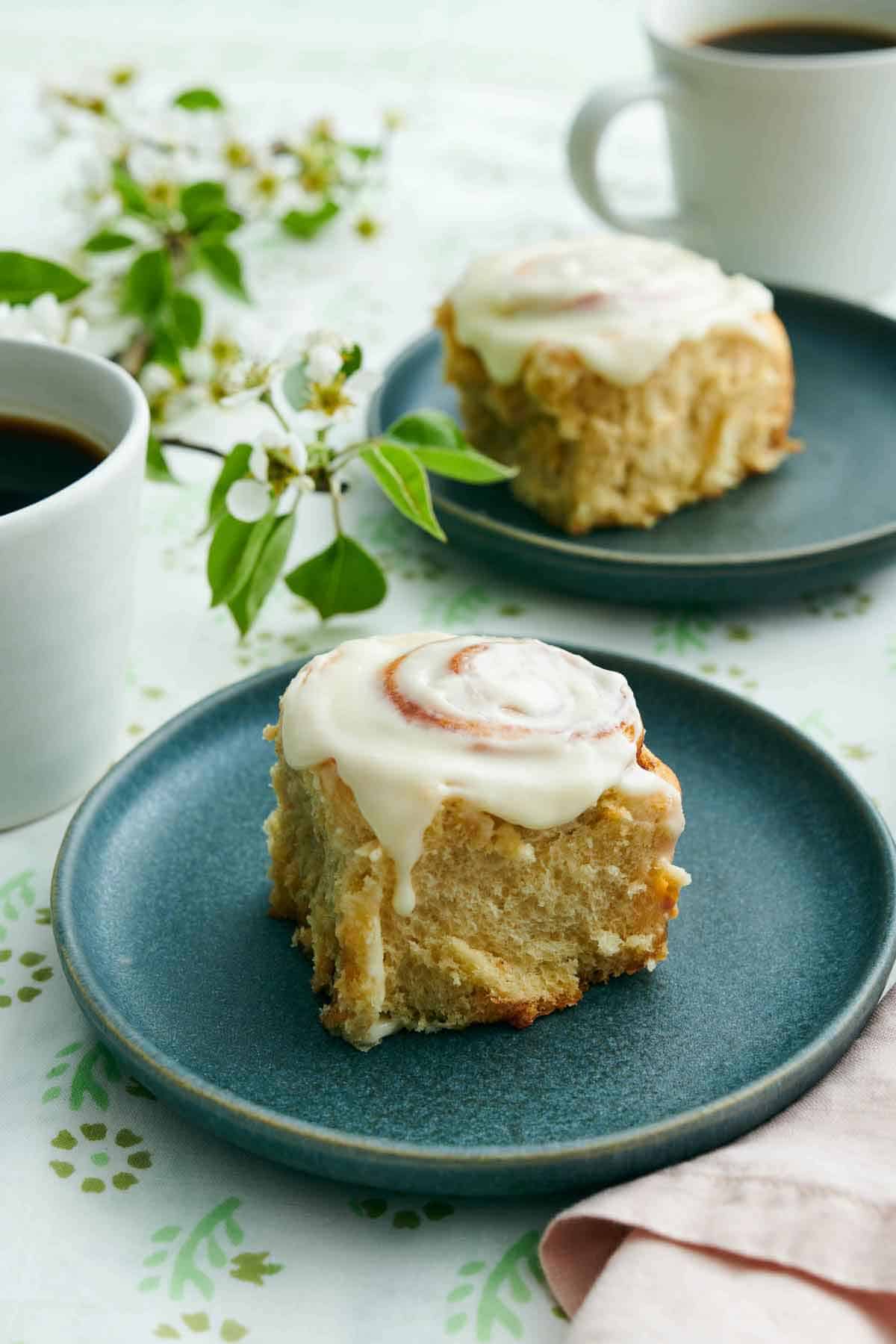 Two plates with cinnamon rolls, one plate in front of the other. Two mugs of coffee placed nearby.