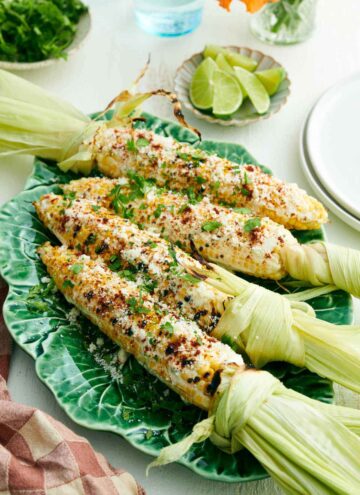 A platter with four elotes with cilantro sprinkled on top. Plate of limes and cilantro in the back.