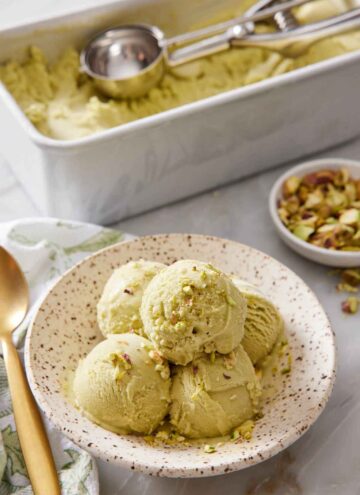 A plate with multiple scoops of pistachio ice cream with more ice cream in the background in a container with an ice cream scoop.