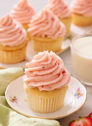 A cupcake topped with strawberry frosting with more topped cupcakes in the background along with a glass of milk.