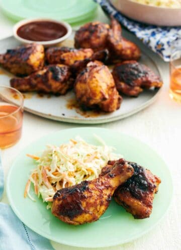 A plate with two pieces of BBQ chicken and coleslaw with a platter of more BBQ chicken in the background.