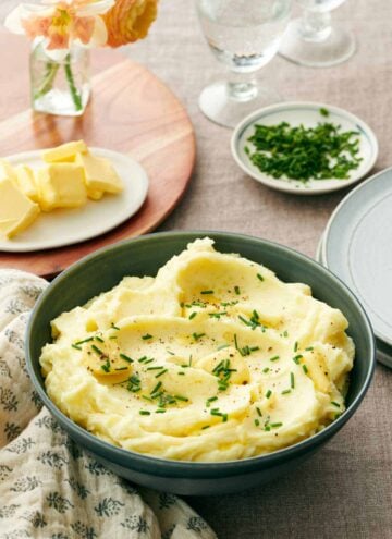 A bowl of garlic mashed potatoes with chives, pepper, and butter on top. More butter in the background, chives, and glasses of water.