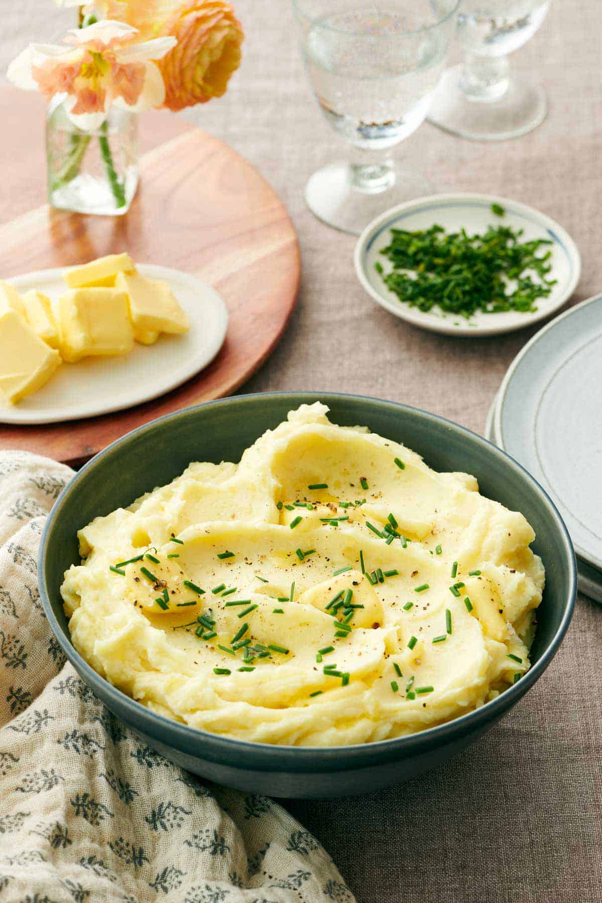 A bowl of garlic mashed potatoes with chives, pepper, and butter on top. More butter in the background, chives, and glasses of water.