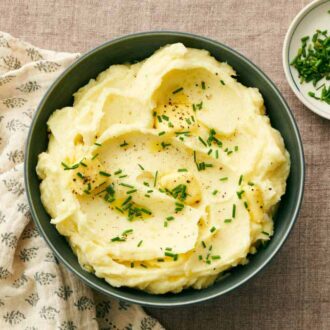 Overhead view of a bowl of garlic mashed potatoes with chives, pepper, and butter with a plate of chives on the side.