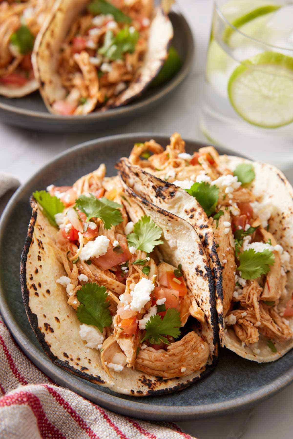 A close up view of two Instant Pot chicken tacos on a plate. A drink with limes in the background and more tacos.