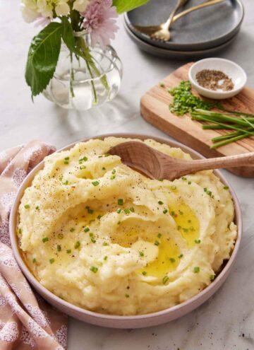 A bowl of Instant Pot mashed potatoes topped with chives and melted butter. A wooden spoon scooped in. Additional toppings and flowers in the background.