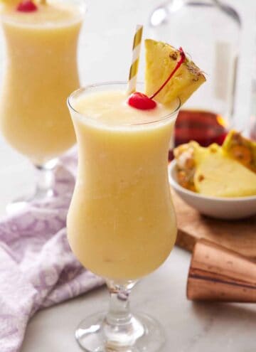 A glass of piña colada with a straw, pineapple slice, and a maraschino cherry. A second glass in the background.