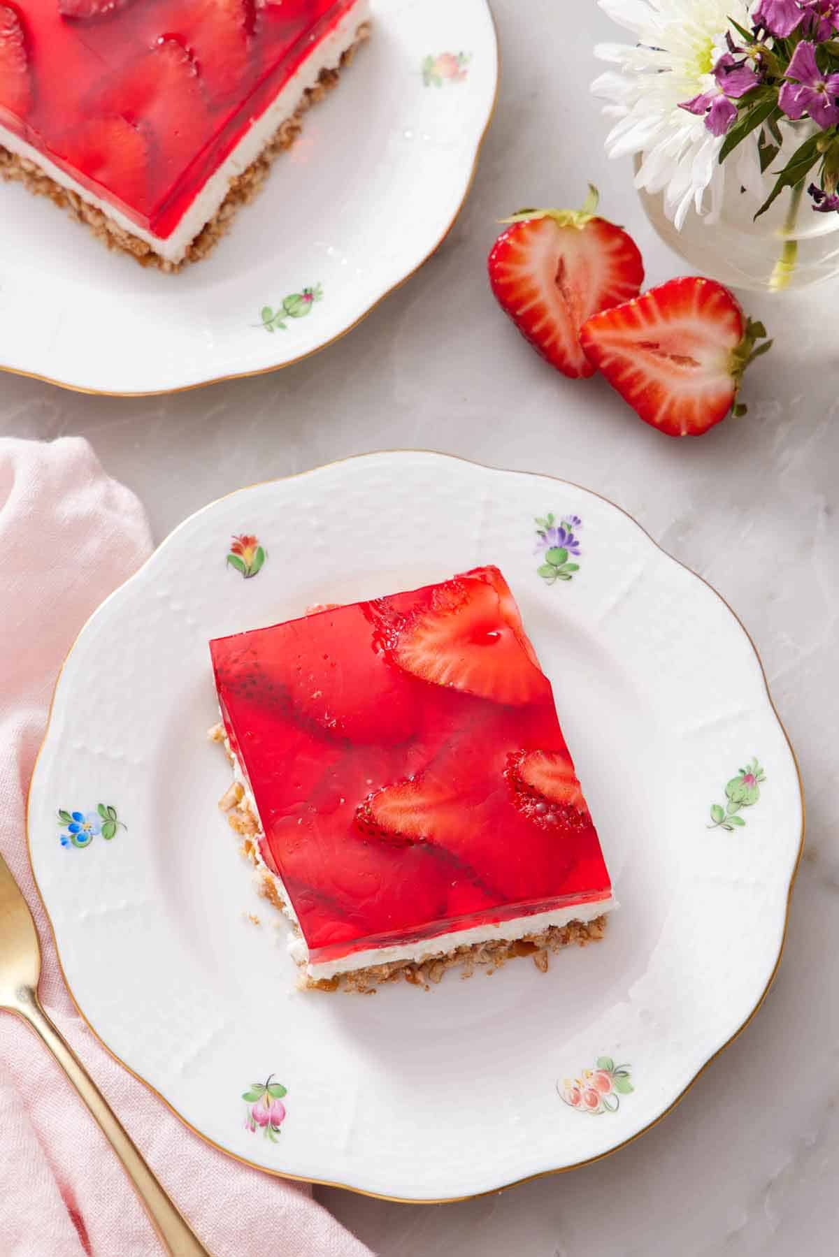 Overhead view of a plate with a square piece of strawberry pretzel salad with cut strawberries, vase of flowers, and another plates slice off to the side.