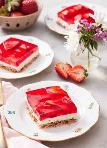 Three plates with sliced square servings of strawberry pretzel salad with a vase of flowers and bowl of strawberries between them.