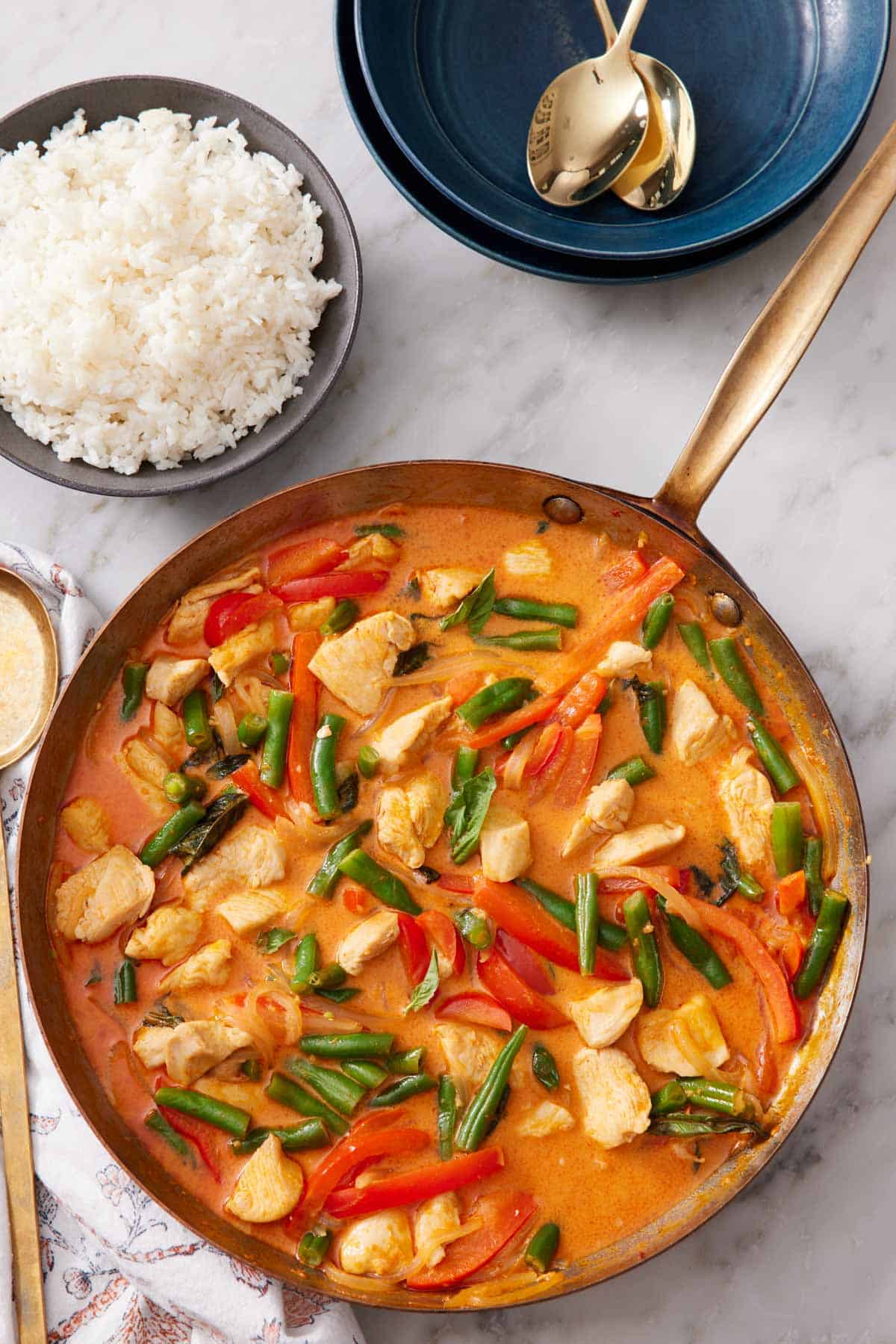 Overhead view of a skillet of Thai red curry with a bowl of rice beside it along with some bowls and spoons.