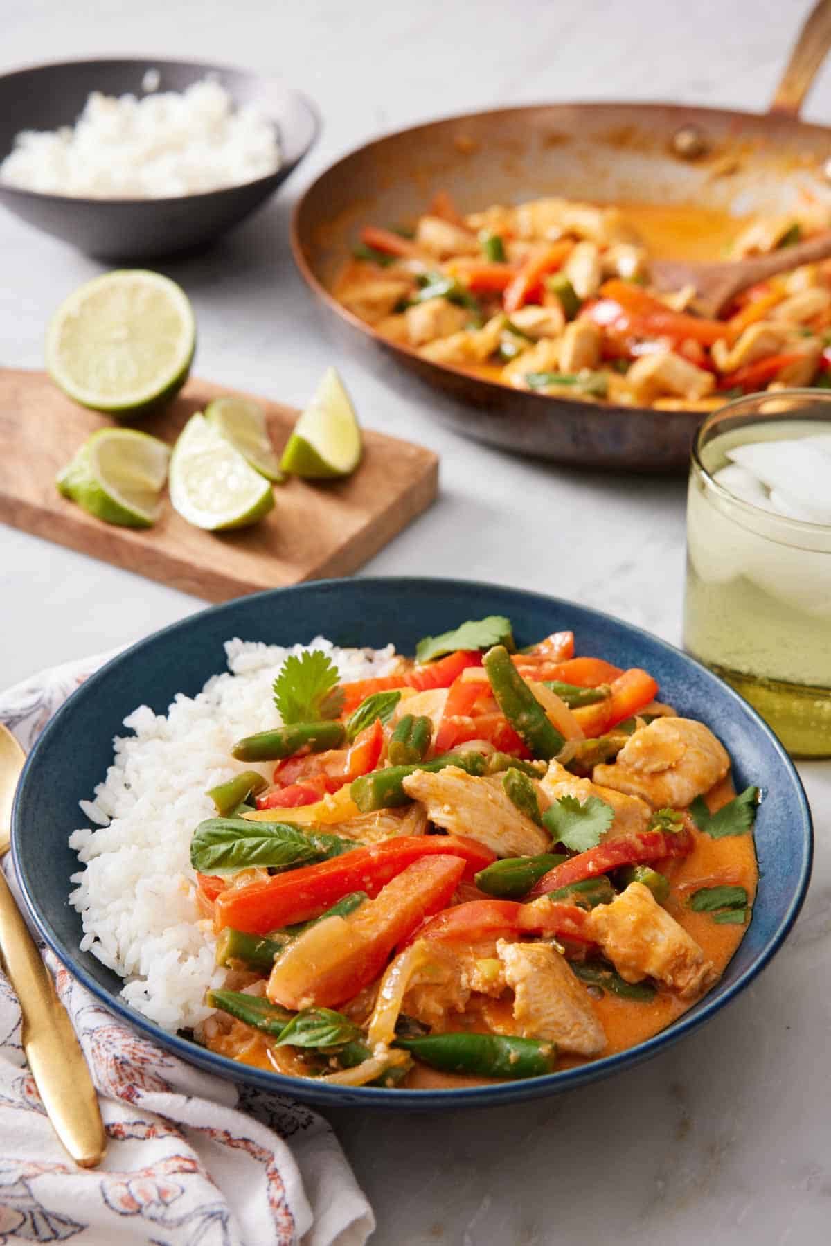 A bowl of Thai red curry with rice. A skillet with more Thai red curry in the background also with limes, a drink, and rice.