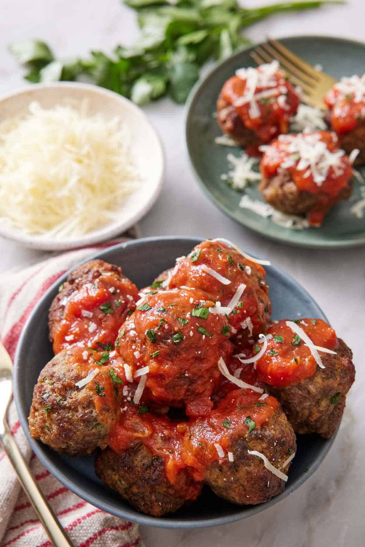 A small bowl of air fryer meatballs with sauce on top along with shredded parmesan. Another plate in the background with a bowl of parmesan.