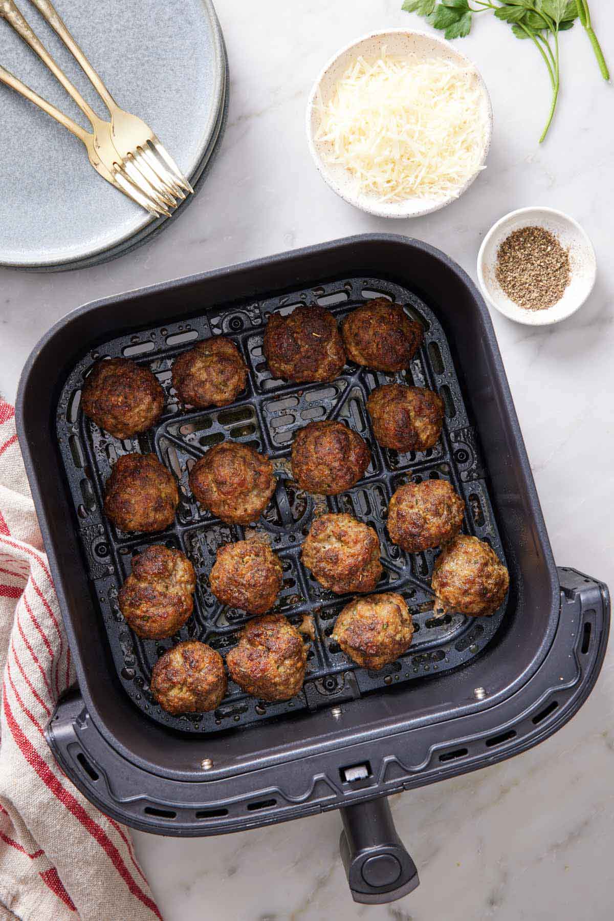Overhead view of air fryer meatballs in an air fryer basket. A bowl of parmesan and a bowl of pepper on the side with a stack of plates and forks.